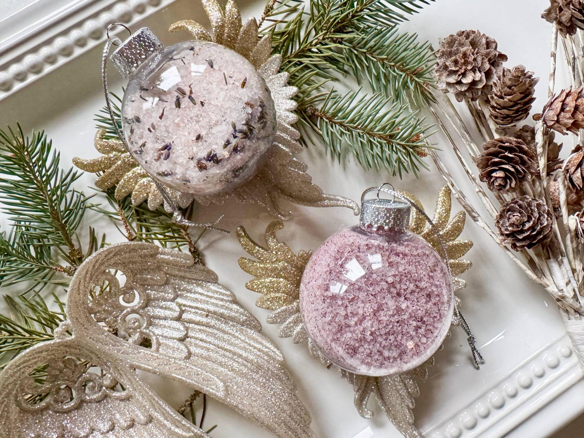 Decorative Christmas ornaments with glittery pink and silver balls, gold angel wings, and pine branches on a white surface.