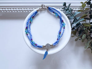 A beaded anklet with light blue, medium blue, and teal beads, featuring a silver-toned circular charm and a blue feather pendant, displayed on a white ceramic dish with green leaves in the background.